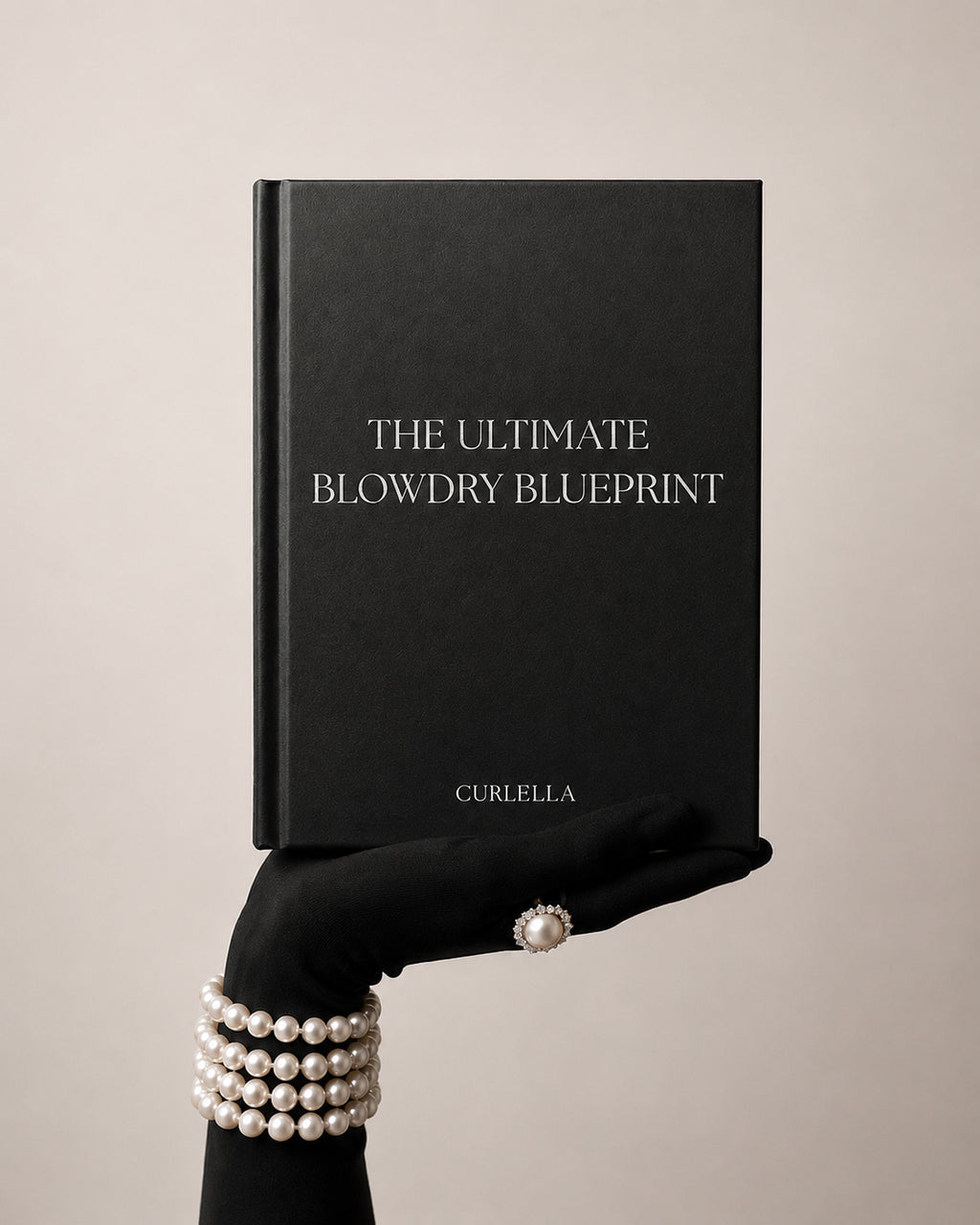 A black hardcover book titled 'THE ULTIMATE BLOWDRY BLUEPRINT' with the brand name 'CURLELLA' at the bottom, held by a gloved hand wearing pearl bracelets and a pearl ring against a warm beige background.
