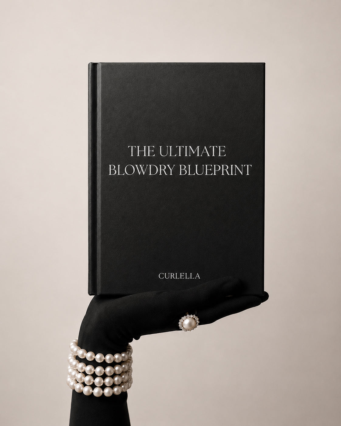 A black hardcover book titled 'THE ULTIMATE BLOWDRY BLUEPRINT' with the brand name 'CURLELLA' at the bottom, held by a gloved hand wearing pearl bracelets and a pearl ring against a warm beige background.