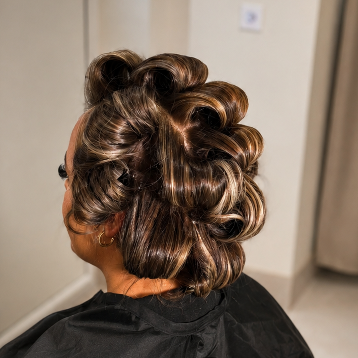 A person seated in a salon chair viewed from the back, wearing a black salon cape. The hair is styled in large, defined curls and waves in dark brown with lighter blonde highlights.