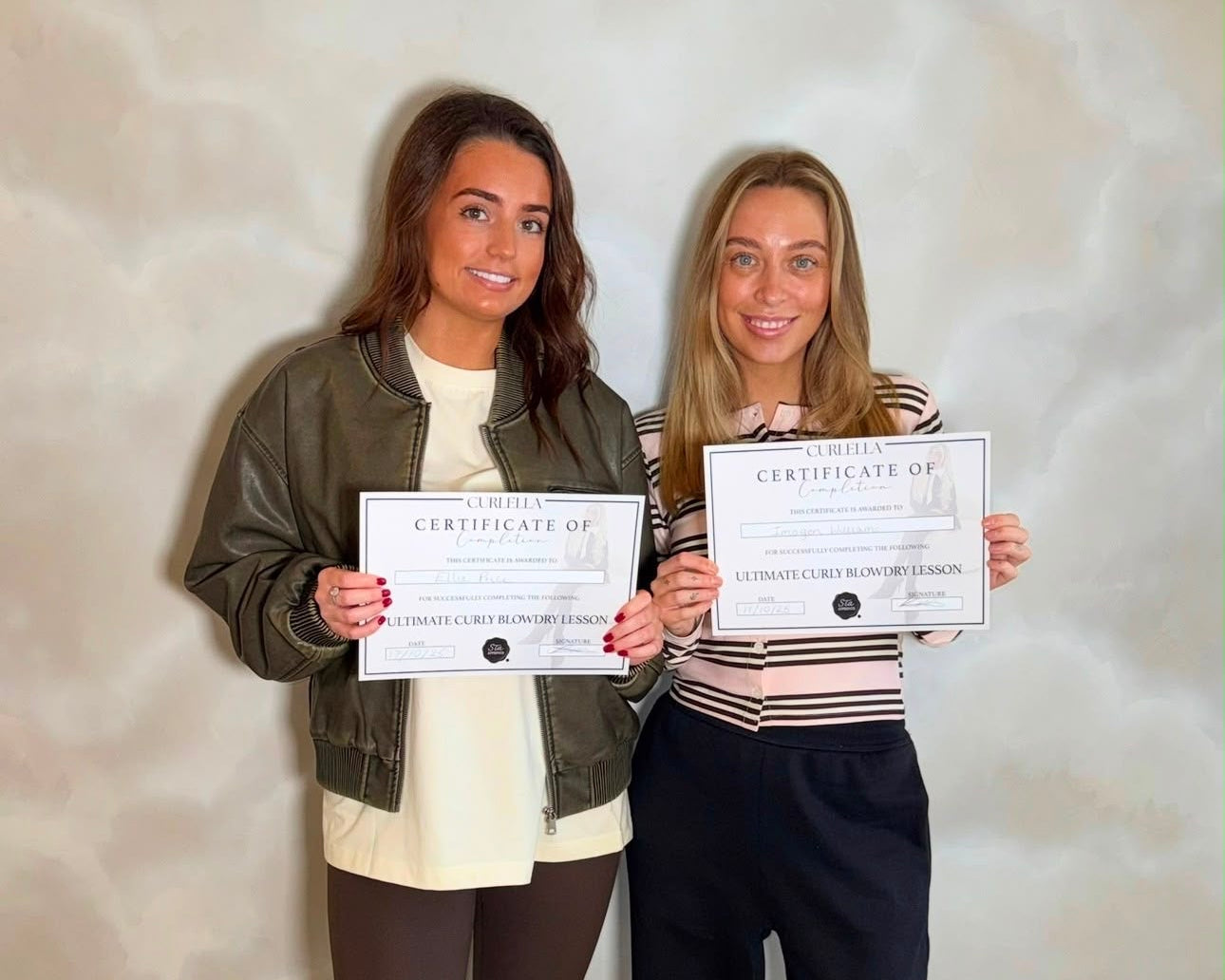 Two women standing indoors and holding printed certificate documents. The woman on the left wears a dark olive jacket over a cream top with dark leggings and white sneakers.