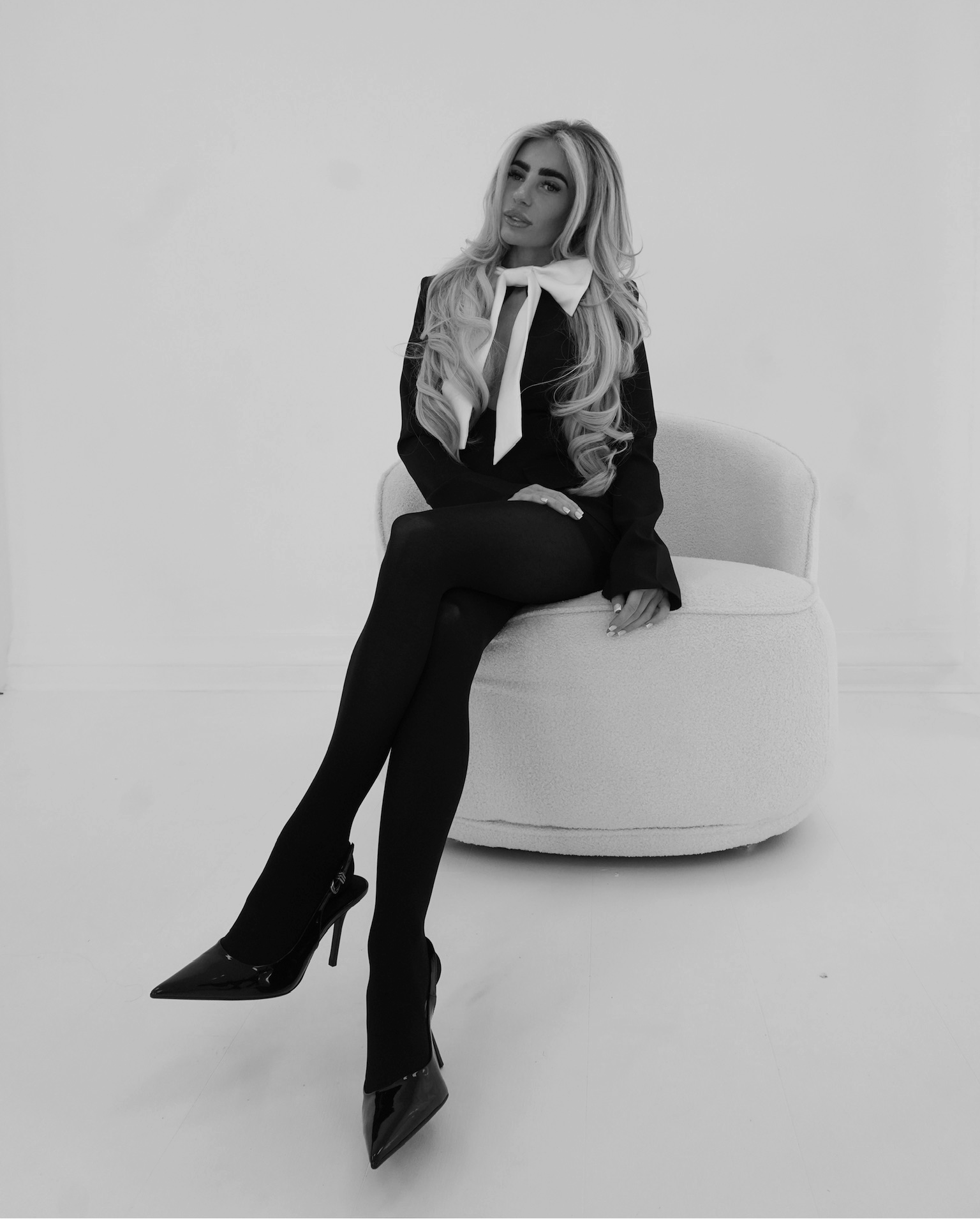 A woman with long blonde hair sits on a white modern chair in a minimalist white studio setting. She is wearing a black blazer over a white collared shirt, a black tie, black tights, and black pointed-toe high heels.