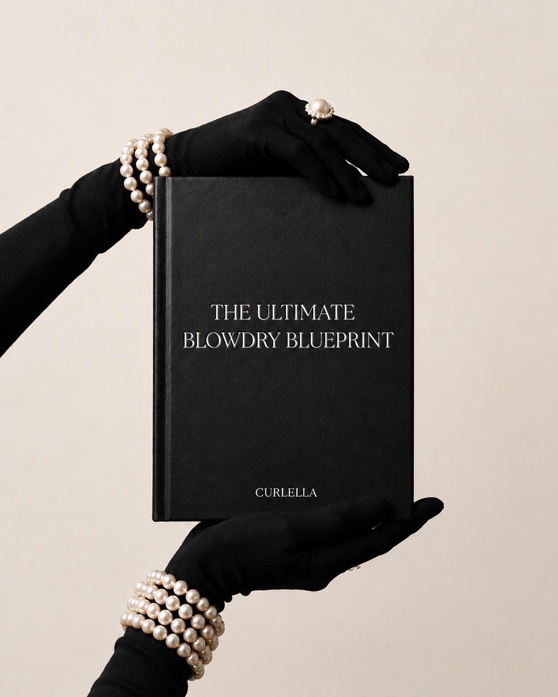 A black hardcover book titled 'THE ULTIMATE BLOWDRY BLUEPRINT' with the brand name 'CURELLA' at the bottom, held by two hands wearing black gloves and pearl bracelets against a warm beige background.