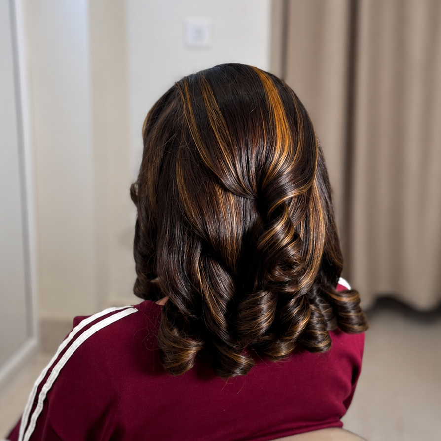 A woman viewed from behind wearing a burgundy top with white stripes on the sleeves, seated in a salon chair. Her hair is styled in loose curls with highlighted brown and golden-blonde streaks.