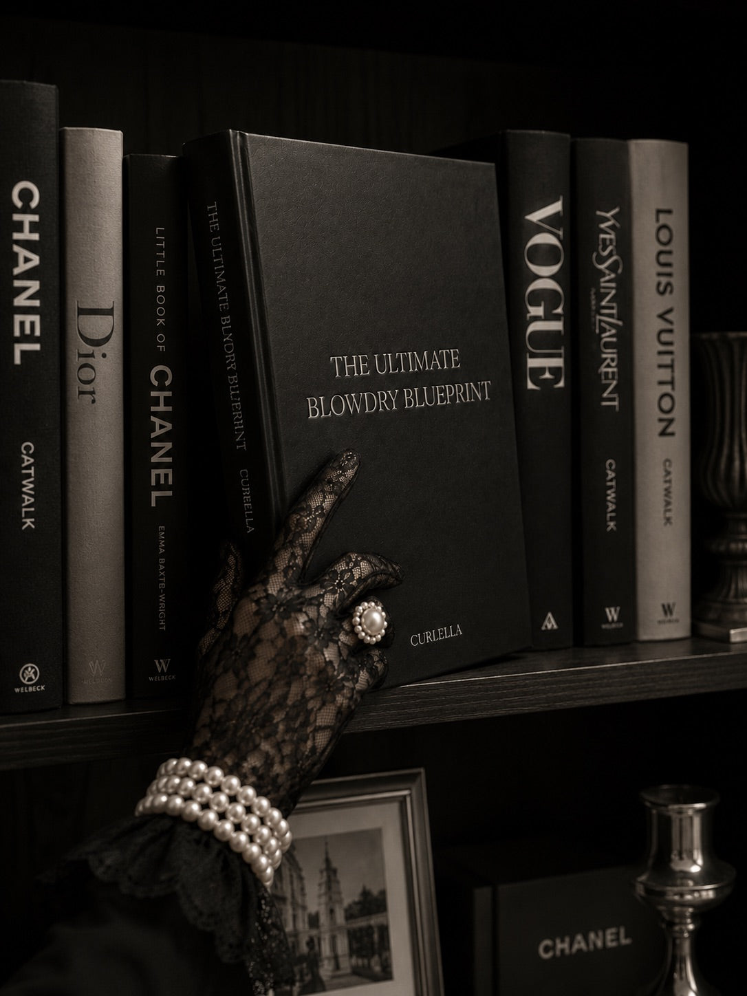 A hand in a black lace glove with pearl bracelets and a statement ring rests on a black book titled 'The Ultimate Blowdry Blueprint' by Criaella, set against a dark bookshelf lined with designer books including Chanel, Dior, Vogue, and Louis Vuitton.