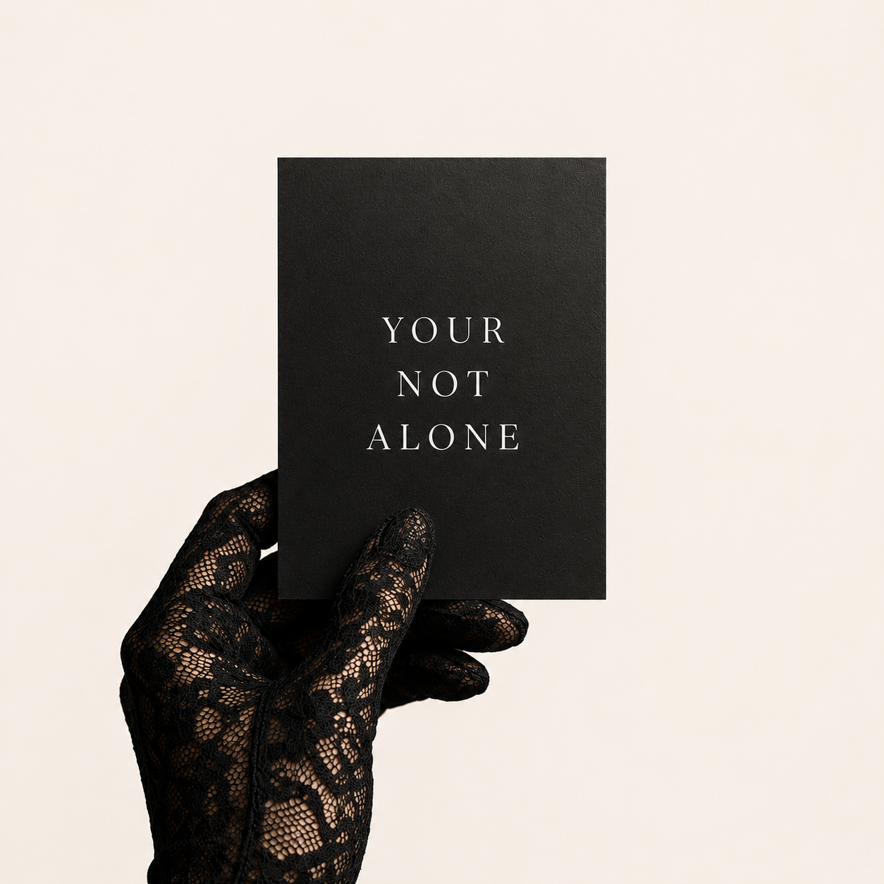 A hand wearing a black lace glove holds a black rectangular card with the words 'YOUR NOT ALONE' printed in white serif text against a plain light beige background.
