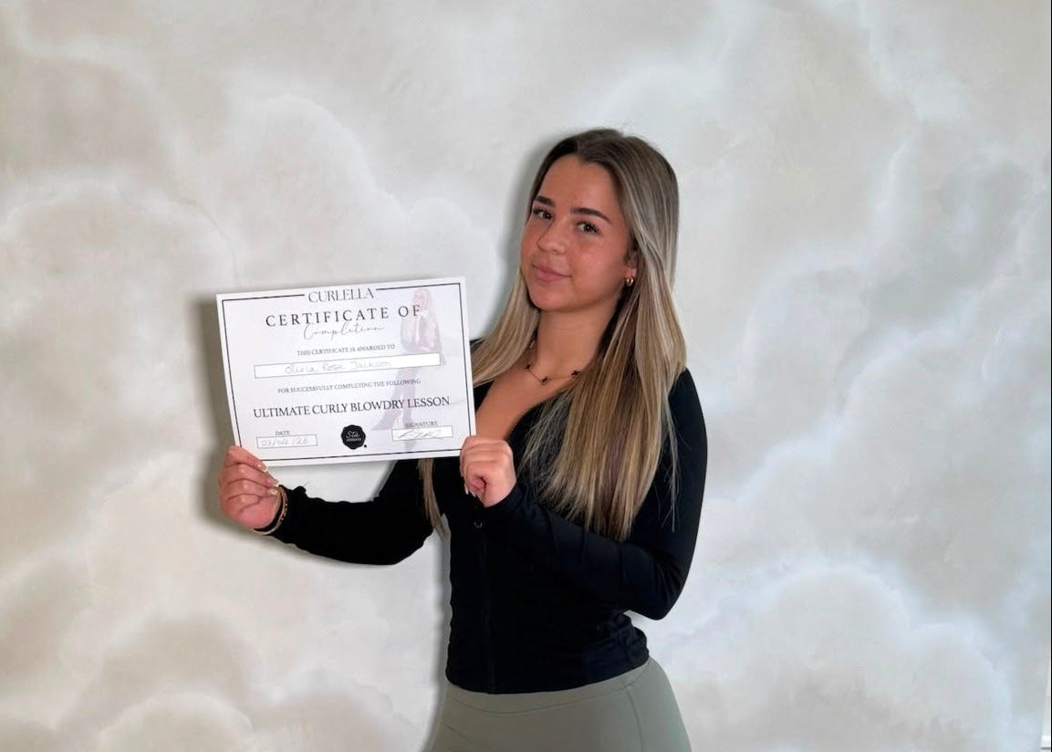 A woman with blonde hair stands indoors against a textured beige wall, holding a framed certificate. She is wearing a black long-sleeve top, olive green pants, and white sneakers.