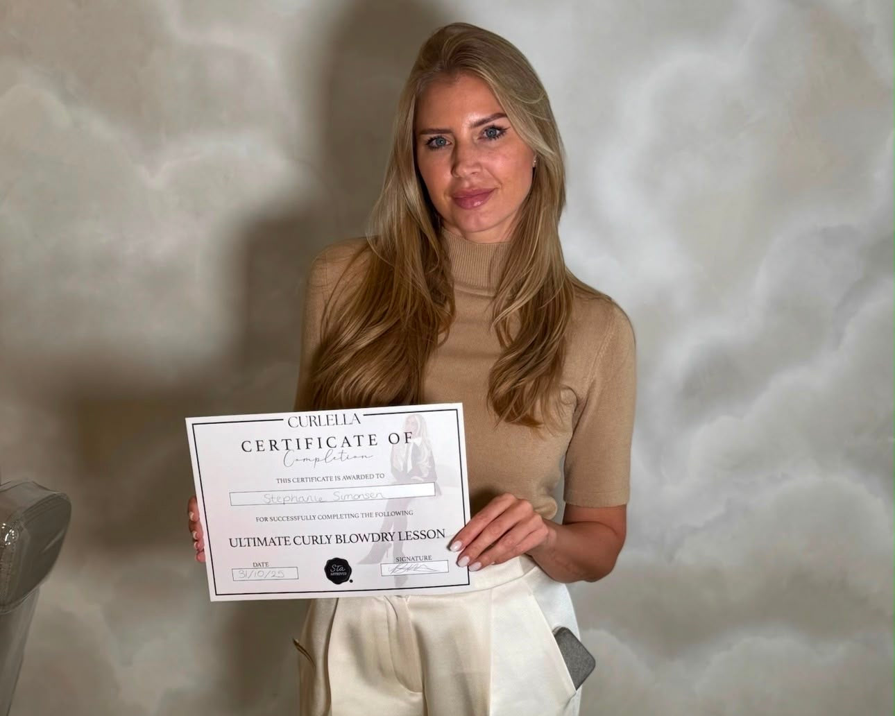 A woman with long blonde hair stands indoors holding a certificate. She is wearing a beige mock-neck top and cream wide-leg trousers.