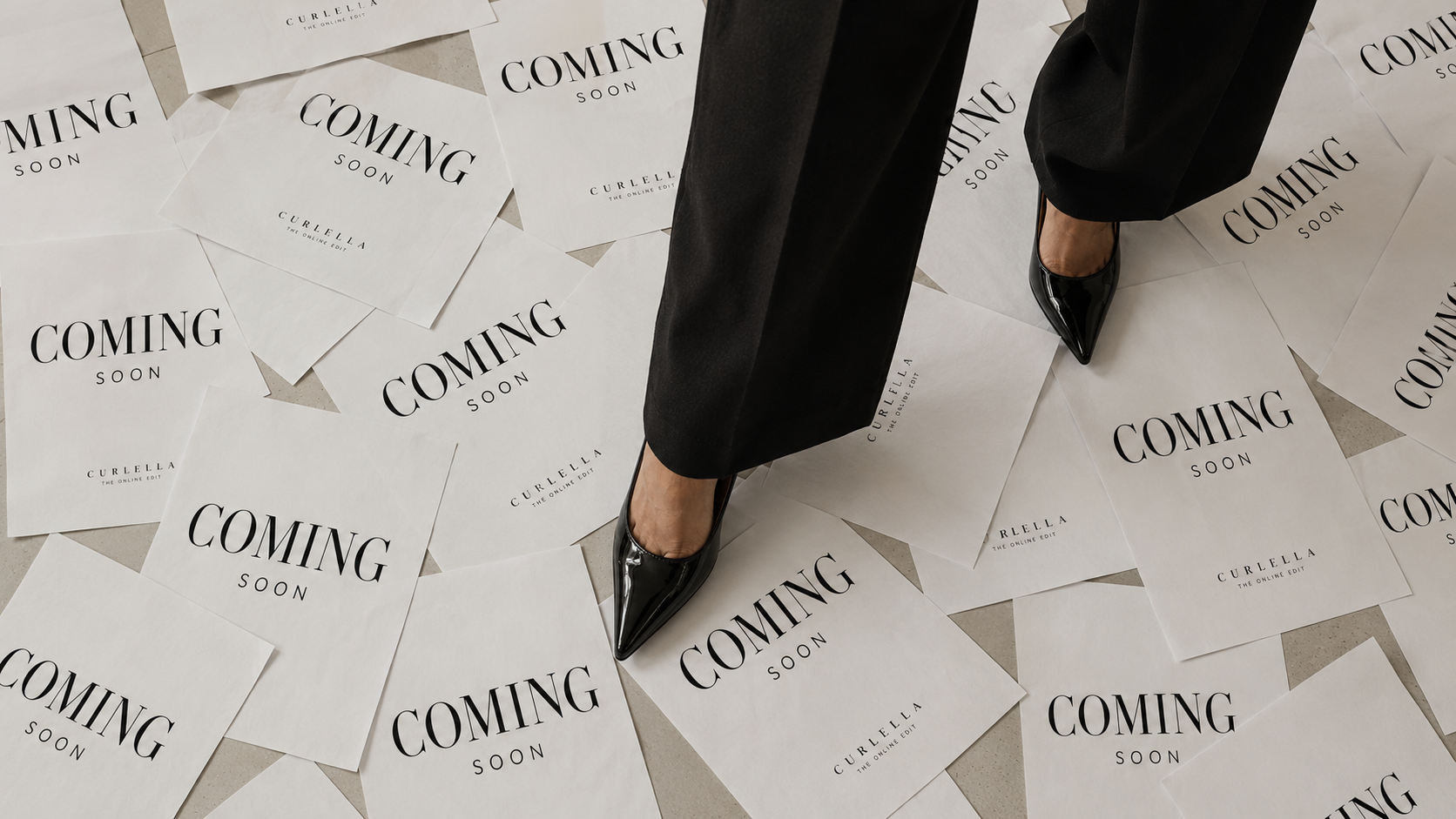 A person wearing black trousers and pointed black heels stands on a floor covered with scattered white cards printed with the words 'COMING SOON'.