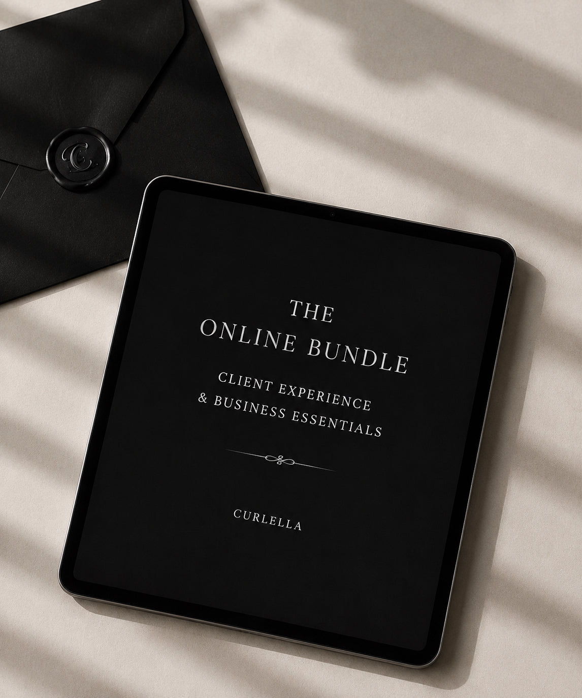 A tablet device displaying a black screen with white text reading 'THE ONLINE BUNDLE', 'CLIENT EXPERIENCE & BUSINESS ESSENTIALS', and 'CURELLA', placed on a light beige surface with soft diagonal shadows. A black envelope with a round wax seal is partially visible in the upper left.