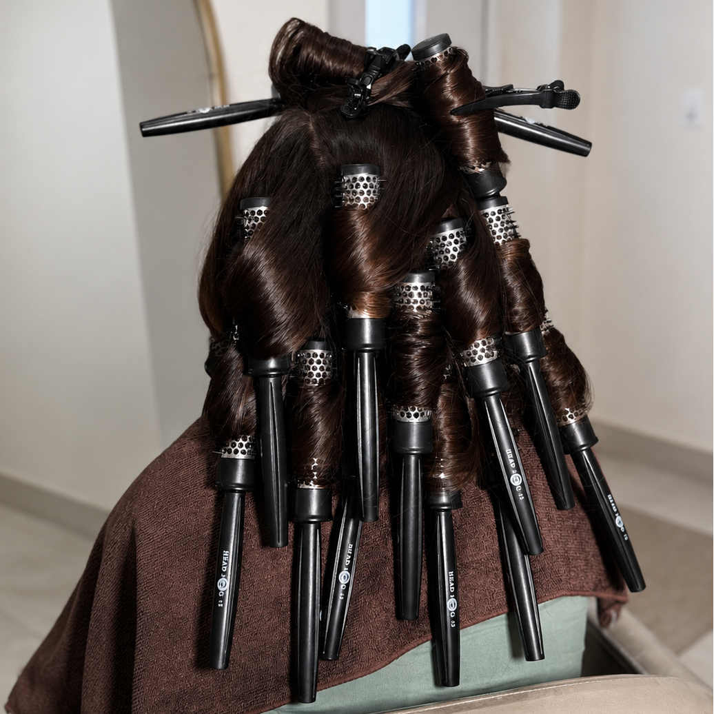 Brown hair styled with multiple black curling irons wrapped in dark brown protective oil, resting on a brown towel in a salon setting.