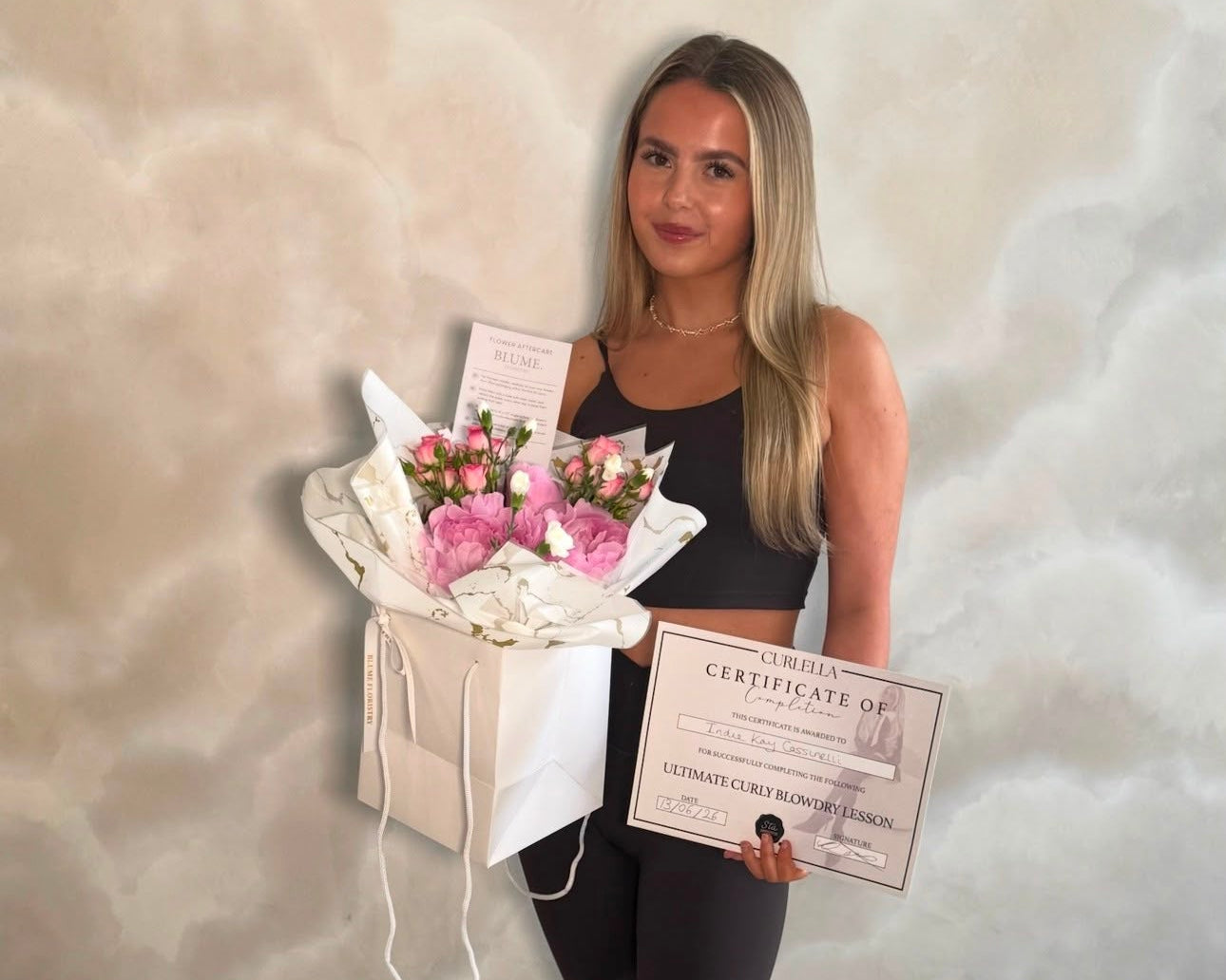 A woman stands indoors holding a bouquet of pink flowers in a white gift bag and a certificate. She is wearing a black tank top, black leggings, and white sneakers against a beige marbled wall.